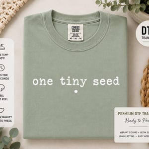 May include: A sage green t-shirt featuring the white text "one tiny seed" printed in a typewriter-style font across the center of the chest. The shirt has a crew neckline and a Comfort Colors brand label visible on the inside of the collar.