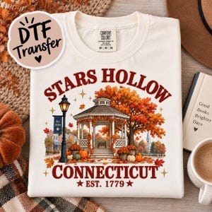May include: A cream-colored t-shirt featuring a graphic of a gazebo in a park during autumn. The text reads Stars Hollow Connecticut Est. 1779. The design includes a street lamp, orange fall foliage, pumpkins, and small stars. The shirt has a Comfort Colors label.