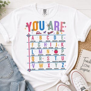Puede incluir: Una camiseta blanca con un gr&aacute;fico colorido que incluye el texto YOU ARE y una tabla alfab&eacute;tica donde cada letra corresponde a una palabra positiva como Accepting, Brave, Creative, Dedicated, Encouraging, Friendly, Genuine, Helpful, Inspiring, Joyful, Kind.