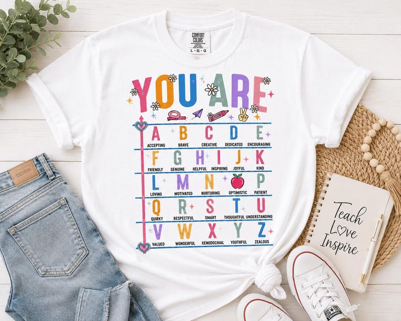 Puede incluir: Una camiseta blanca con un gr&aacute;fico colorido que incluye el texto YOU ARE y una tabla alfab&eacute;tica donde cada letra corresponde a una palabra positiva como Accepting, Brave, Creative, Dedicated, Encouraging, Friendly, Genuine, Helpful, Inspiring, Joyful, Kind.