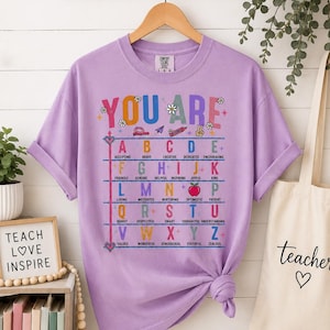 Puede incluir: Una camiseta de manga corta color lavanda con un estampado gr&aacute;fico colorido. El texto YOU ARE aparece sobre una tabla alfab&eacute;tica donde cada letra corresponde a un adjetivo positivo. Incluye peque&ntilde;os iconos como un l&aacute;piz, una manzana y flores.