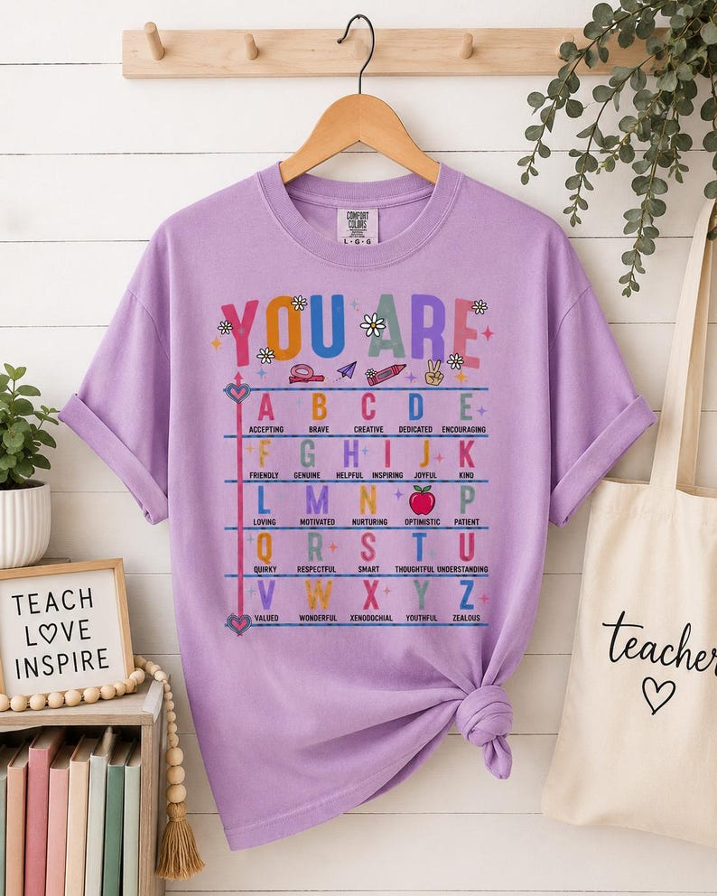 Puede incluir: Una camiseta de manga corta color lavanda con un estampado gr&aacute;fico colorido. El texto YOU ARE aparece sobre una tabla alfab&eacute;tica donde cada letra corresponde a un adjetivo positivo. Incluye peque&ntilde;os iconos como un l&aacute;piz, una manzana y flores.