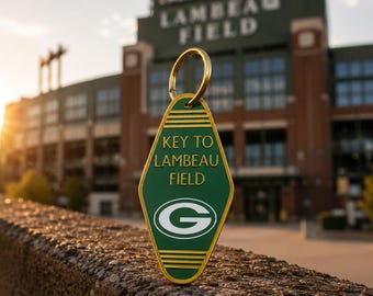 May include: A diamond-shaped green hotel-style keychain with gold trim and a gold metal ring. The text reads KEY TO LAMBEAU FIELD in gold lettering above a white and green G logo. The keychain features horizontal gold lines at the top and bottom.