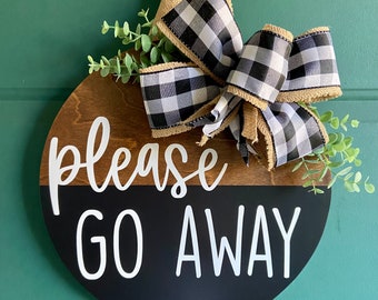 May include: A round wooden sign with a black and white checkered bow and greenery. The sign says "Please Go Away" in white lettering.