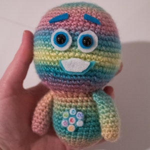 Può includere: Un giocattolo di peluche all'uncinetto color arcobaleno con un sopracciglio di feltro blu, occhi di feltro blu e una bocca di feltro bianco. Il giocattolo ha un design circolare sul petto.