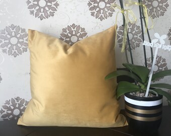 May include: A mustard yellow velvet pillow with a white background featuring a floral pattern. The pillow is sitting on a brown surface.