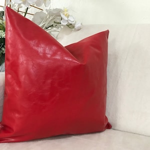 May include: A vibrant red square pillow with a glossy, leather-like texture. The pillow is placed on a white sofa, with white flowers in the background. This decorative pillow adds a pop of color to any living space.