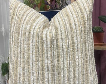 Puede incluir: Un cojín decorativo cuadrado con un patrón de rayas texturizadas en tonos beige y crema. El cojín está colocado sobre una superficie azul oscuro, con plantas y una estantería en el fondo. El cojín tiene un aspecto suave y acogedor.