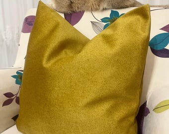 May include: A golden-yellow decorative pillow with a textured surface rests on a floral patterned sofa. A golden-brown cat is perched on the sofa, above the pillow. The sofa has a white background with purple, teal, and green floral designs.