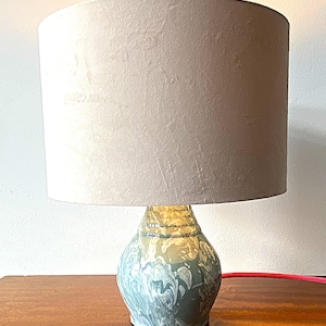 May include: A decorative table lamp with a light gray lampshade and a marbled blue and white ceramic base. The lamp has a cylindrical shade and a bulbous base. The lamp is on a wooden surface.