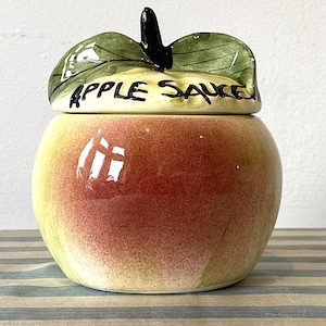 Può includere: Un barattolo in ceramica a forma di mela con coperchio. Il barattolo è dipinto con un gradiente di giallo e rosso, che ricorda una mela matura. Il coperchio è verde con un gambo nero e le parole "APPLE SAUCE" sono scritte sul coperchio.