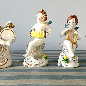 Puede incluir: Conjunto de cuatro figuras de querubines de cerámica, cada una tocando un instrumento musical diferente. Los querubines tienen cabello castaño, alas azules y doradas, y están sentados sobre bases blancas ornamentadas con detalles dorados y flores amarillas.