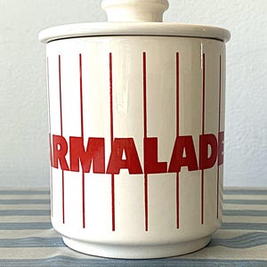 Può includere: Un barattolo di marmellata in ceramica bianca con coperchio. Il barattolo presenta strisce rosse verticali e la scritta "MARMALADE" in lettere rosse. Il barattolo è su una superficie a righe blu e bianche.
