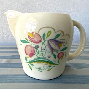 Könnte beinhalten: Cremefarbener Keramikkrug mit floralem Muster. Der Krug hat einen Ausguss und einen Henkel. Das florale Design zeigt rosa, lila und gelbe Blumen mit grünen Blättern und Stielen. Der Krug steht auf einer blau-weiß gestreiften Oberfläche.