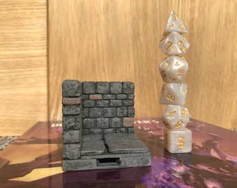 May include: A white stone dice tower with a gray stone wall design. The tower has a small opening at the bottom. A stack of six white dice with gold numbers is next to the tower.