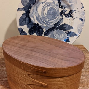 Puede incluir: Una caja ovalada de madera con tapa con bisagras y detalles decorativos de madera. La caja está hecha de madera marrón claro y tiene un acabado liso. La caja está sobre una superficie de madera.