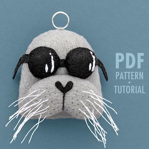 May include: A gray felt seal head with black sunglasses and white whiskers. The seal has a black heart-shaped nose and a silver ring at the top. Text on the right reads "PDF PATTERN + TUTORIAL".