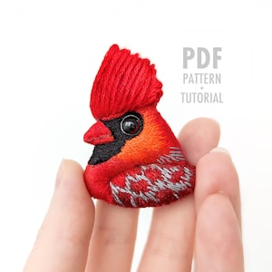 Puede incluir: Broche bordado de un cardenal con detalles rojos, naranjas, negros y grises. El pájaro tiene una cresta prominente y está sostenido por una mano. La imagen incluye el texto "PDF PATTERN + TUTORIAL".