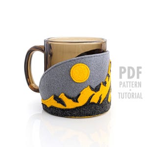 May include: A clear glass mug with a felt cozy featuring a mountain landscape design. The cozy is gray with yellow mountains and a yellow sun. The text "PDF PATTERN + TUTORIAL" is visible.