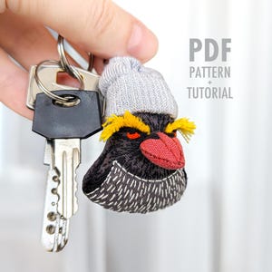 May include: Embroidered penguin keychain with a gray beanie, yellow accents, and a red beak. The keychain is attached to a set of keys. Text on the right reads "PDF PATTERN + TUTORIAL".