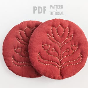 May include: Two round, red fabric coasters with embroidered floral designs. The coasters have a quilted texture and are adorned with beige stitching. The text "PDF PATTERN + TUTORIAL" is in the upper left corner.