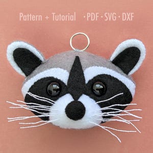 May include: A handmade raccoon ornament with a gray and black face, white accents, and a metal ring for hanging. The text "Pattern + Tutorial PDF SVG DXF" is at the top.