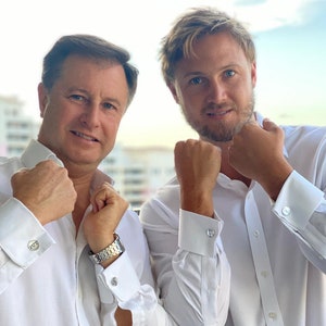 Puede incluir: Dos hombres con camisas blancas de vestir con gemelos. Los hombres están de pie uno al lado del otro y sonriendo a la cámara.