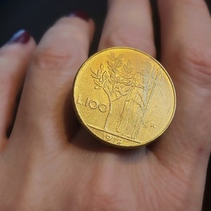 May include: A gold-colored coin ring featuring a woman standing next to a tree with the year 1979 and the number 100 inscribed on the ring.