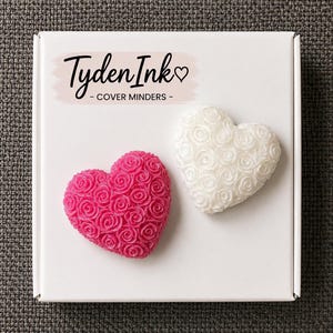 May include: A white square box featuring the text Tyden Ink COVER MINDERS with a heart symbol. Inside the box are two heart-shaped objects, one bright pink and one white, both covered in a textured pattern of small, sculpted rose blossoms.
