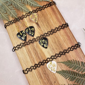 Peut inclure: Trois colliers ras du cou noirs avec des breloques en forme de médiator de guitare dorées. Les breloques sont ornées de différentes notes de musique. Les colliers sont posés sur une surface en bois.