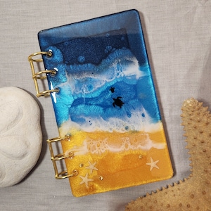 May include: A blue, white, and gold resin notebook with a beach scene and sea turtles. The notebook has gold rings and is closed.