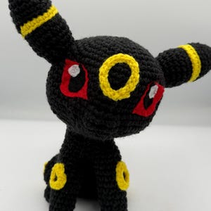 May include: A handmade crochet Umbreon plushie. The plushie is primarily black with yellow accents on the ears, legs, and tail. It has red eyes with white highlights and a yellow ring on its forehead.