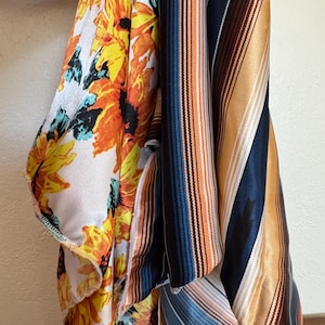 Op de afbeelding: Assortiment van gedrapeerde stoffen met levendige patronen. Eén stof toont grote gele en oranje zonnebloemen op een witte achtergrond. Andere stoffen tonen verticale strepen in tinten blauw, oranje en bruin.