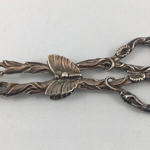 May include: Silver butterfly-shaped sugar tongs with ornate floral details. The tongs are made of silver and have a delicate design.