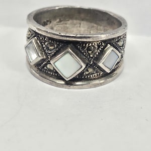 May include: A wide, silver-toned ring featuring three square, iridescent mother-of-pearl inlays. The ring is decorated with small, round, dark accents, possibly marcasite stones, and has a slightly tarnished appearance, suggesting an antique or vintage style.