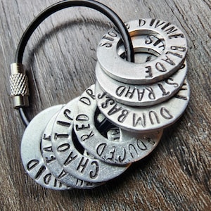 May include: A set of silver keychains with different words engraved on them, including "DUD", "RAHD", "BAST", "DR", "CHA", "RED", "LOVE", "DID". The keychains are attached to a black cord.