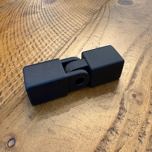 May include: A matte black articulated connector piece featuring two rectangular end blocks joined by a central hinge mechanism with a circular opening. The object has a textured surface finish and a geometric design with clean, sharp edges throughout.