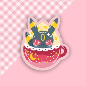 以下が含まれることがあります： 三日月と星のデザインが施された赤とピンクのマグカップ。片目のある黒と黄色の漫画の生き物がマグカップの中にいます。