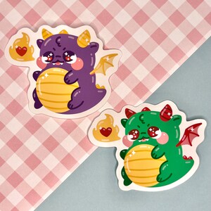Puede incluir: Dos pegatinas de dragones de dibujos animados con corazones y alas. El drag&oacute;n morado sostiene una bola amarilla y el drag&oacute;n verde sostiene una bola amarilla. Ambos dragones tienen corazones rojos en las mejillas.