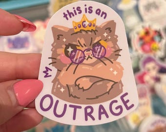 Peut inclure: Un autocollant découpé représentant un chat brun à l'air grincheux portant une couronne jaune et des lunettes rondes violettes. Le chat a les bras croisés, le texte au-dessus indique this is an et le texte en dessous indique OUTRAGE.