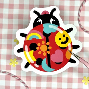 May include: A red ladybug sticker with a black outline and various colorful patterns on its back, including a rainbow, a sun, a heart, a star, and a crescent moon.