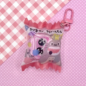 Puede incluir: Un llavero de plástico rosa y transparente con forma de bolsa de caramelo con el texto "sugar sprites" impreso en él. La bolsa contiene un pequeño personaje de dibujos animados gris y blanco con una corona y una mariposa rosa.