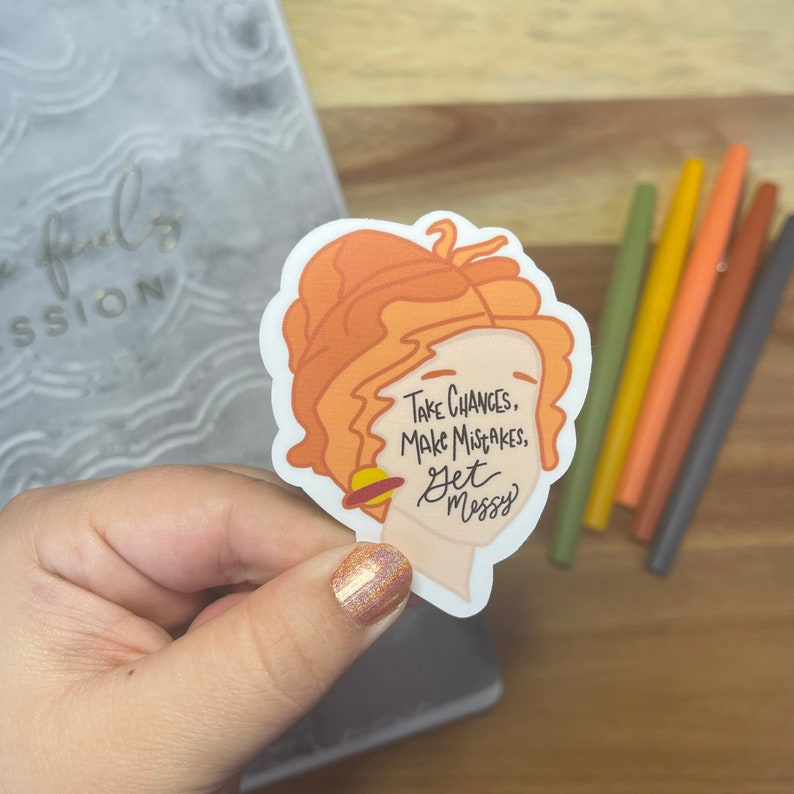 May include: A sticker with a cartoon illustration of a woman with orange hair and a planet around her neck. The sticker has the text "Take chances, make mistakes, get messy".