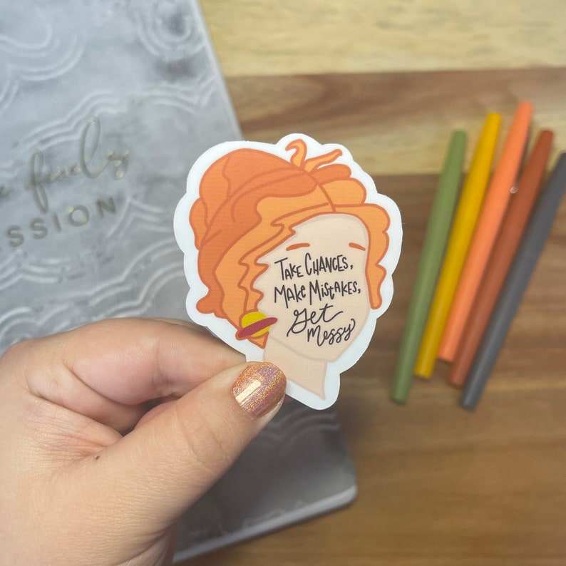 May include: A sticker featuring a cartoon illustration of a woman with orange hair and a planet behind her head. The text on the sticker reads "Take chances, make mistakes, get messy."