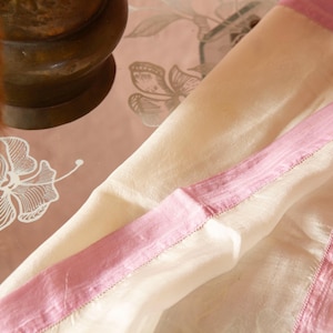 May include: A cream-coloured fabric with a pink border, possibly a scarf or shawl, is draped over a floral patterned surface. The fabric has a soft, flowing appearance. A bronze vase with dried plants is in the background.