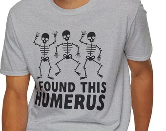 Può includere: T-shirt grigio con grafica nera di tre scheletri che ballano. Gli scheletri alzano le braccia. Il testo "FOUND THIS HUMERUS" è stampato in nero sotto gli scheletri.