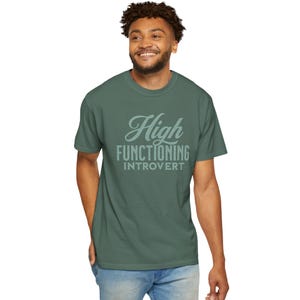 Könnte beinhalten: Ein grünes T-Shirt mit dem Text "High Functioning Introvert" in hellgrüner, stilisierter Schrift. Das Shirt ist einfarbig und hat kurze Ärmel.