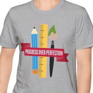 Könnte beinhalten: Ein graues T-Shirt mit einer Bleistift-, Lineal- und Pinselgrafik und dem Text "PROGRESS OVER PERFECTION" auf einem roten Banner. Das T-Shirt wird von einer Person in blauen Jeans getragen.