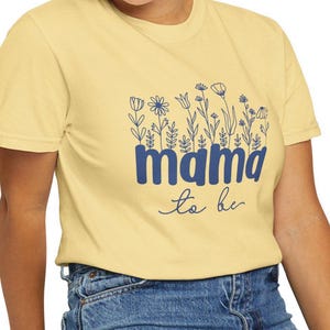 May include: A light yellow t-shirt with the text "mama to be" in blue, adorned with floral illustrations. The shirt is paired with blue jeans.