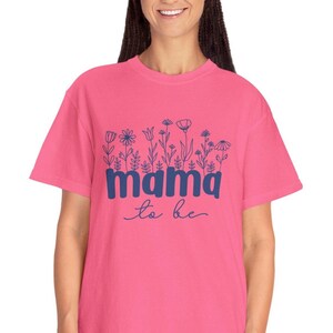 May include: A pink t-shirt with the words "mama to be" in blue script. The word "mama" is in a bold, blocky font, and the shirt features a floral design above the text. The shirt is a short-sleeved, crew neck style.